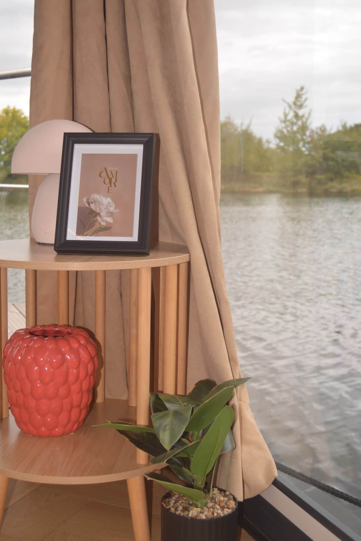 Una habitación con vista a un cuerpo de agua, un cuadro enmarcado, un florero rojo y una planta en maceta sobre una mesa de madera.