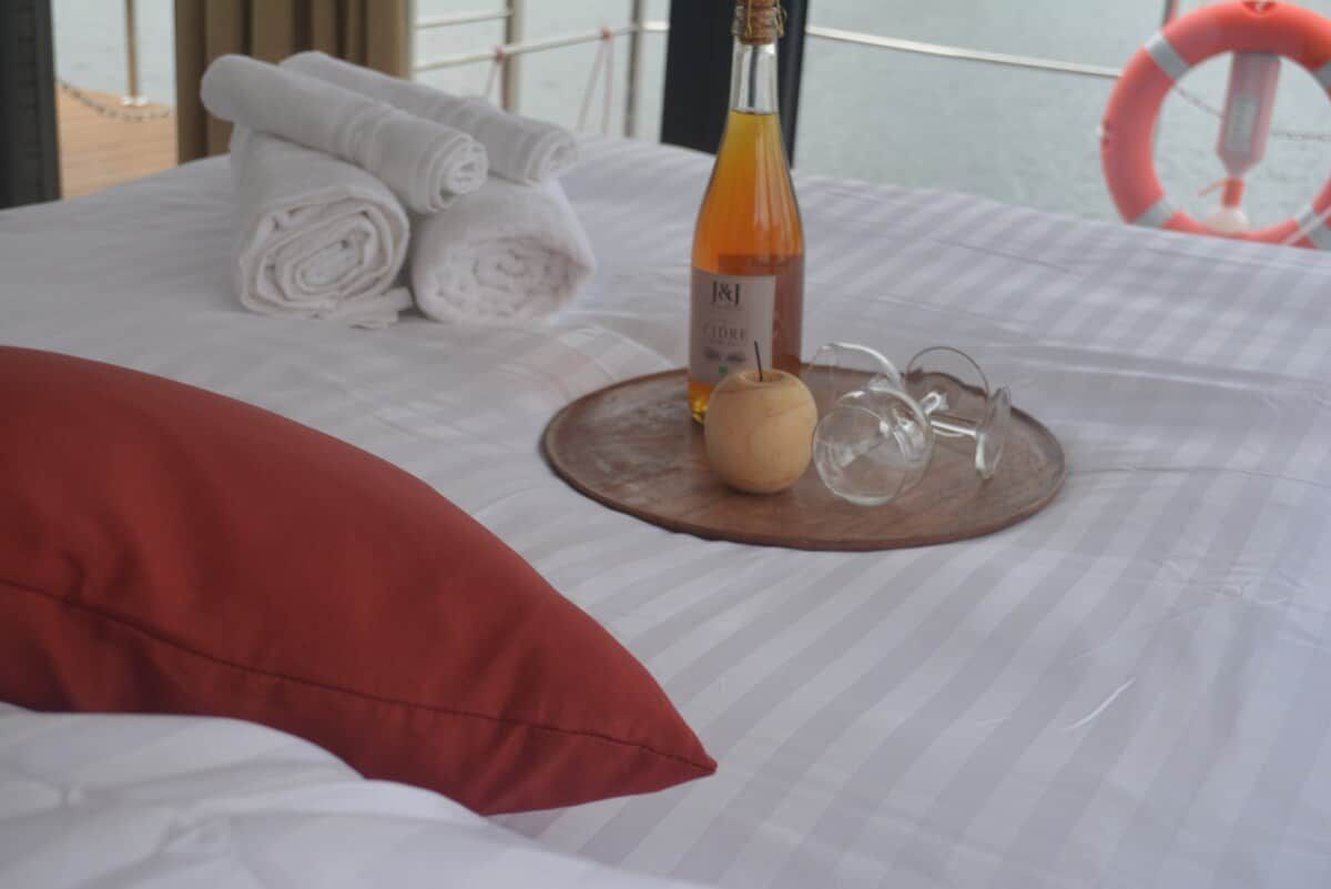 A neatly made bed with rolled towels, a bottle of wine, and a pair of glasses on a wooden tray.