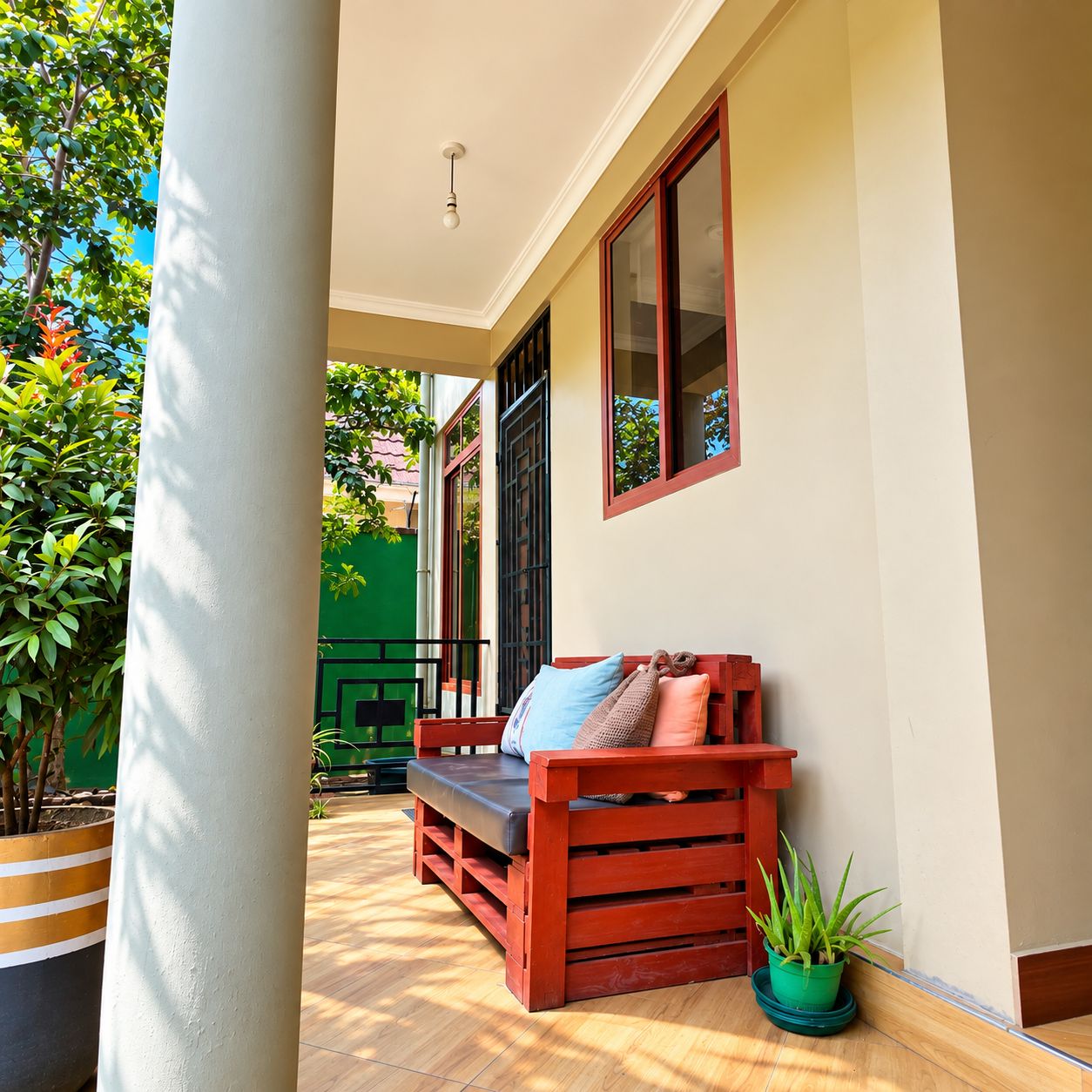 A wooden bench with cushions, a potted plant, and a green wall in a sunlit outdoor area.