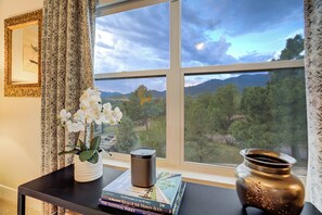A room with a view of mountains through a window, a table with books and a speaker, and a vase with flowers. - Red Rock Retreat | Hot Tub, EV Charger & Office (Colorado Springs)
