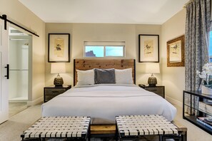 A neatly arranged bedroom with a large bed, two bedside tables with lamps, a window with a view, and a door leading to another room. - Red Rock Retreat | Hot Tub, EV Charger & Office (Colorado Springs)