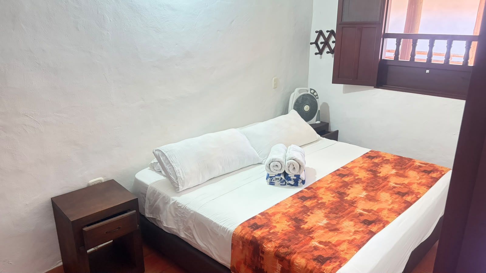 A single bed with white bedding, a small bedside table, a fan, and a window with a view.
