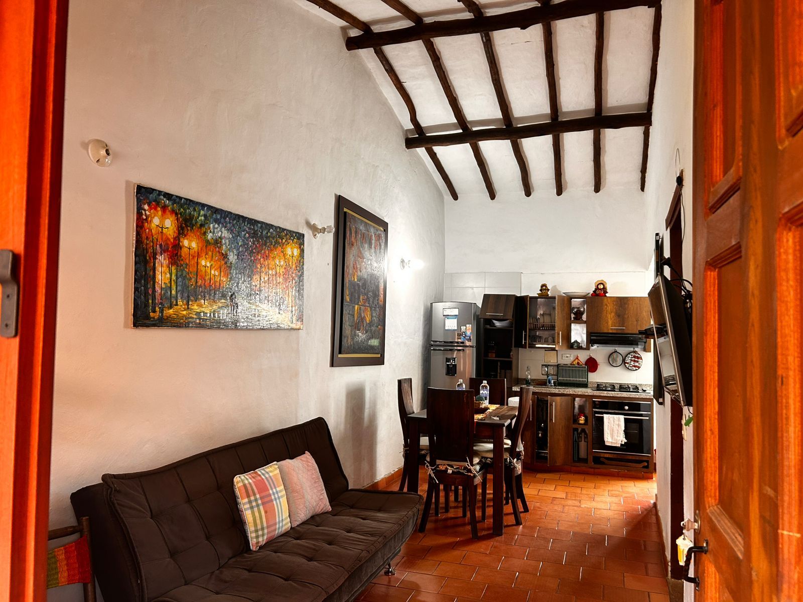 A cozy living room with a sofa and a painting, a kitchen with modern appliances, and a dining area with chairs.