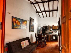 Un saló acollidor amb un sofà i un quadre, una cuina amb electrodomèstics moderns, i una zona de menjador amb cadires.