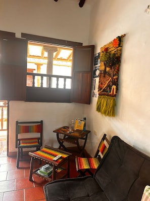Una habitación acogedora con un sofá negro, una mesa de madera con una silla, un ventanal con vistas a un edificio y una pared decorada con un tapiz.