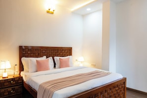 Une chambre d’hôtel avec une tête de lit en bois, un grand lit avec une literie blanche et rose, deux tables de chevet avec des lampes et un luminaire fixé au mur.