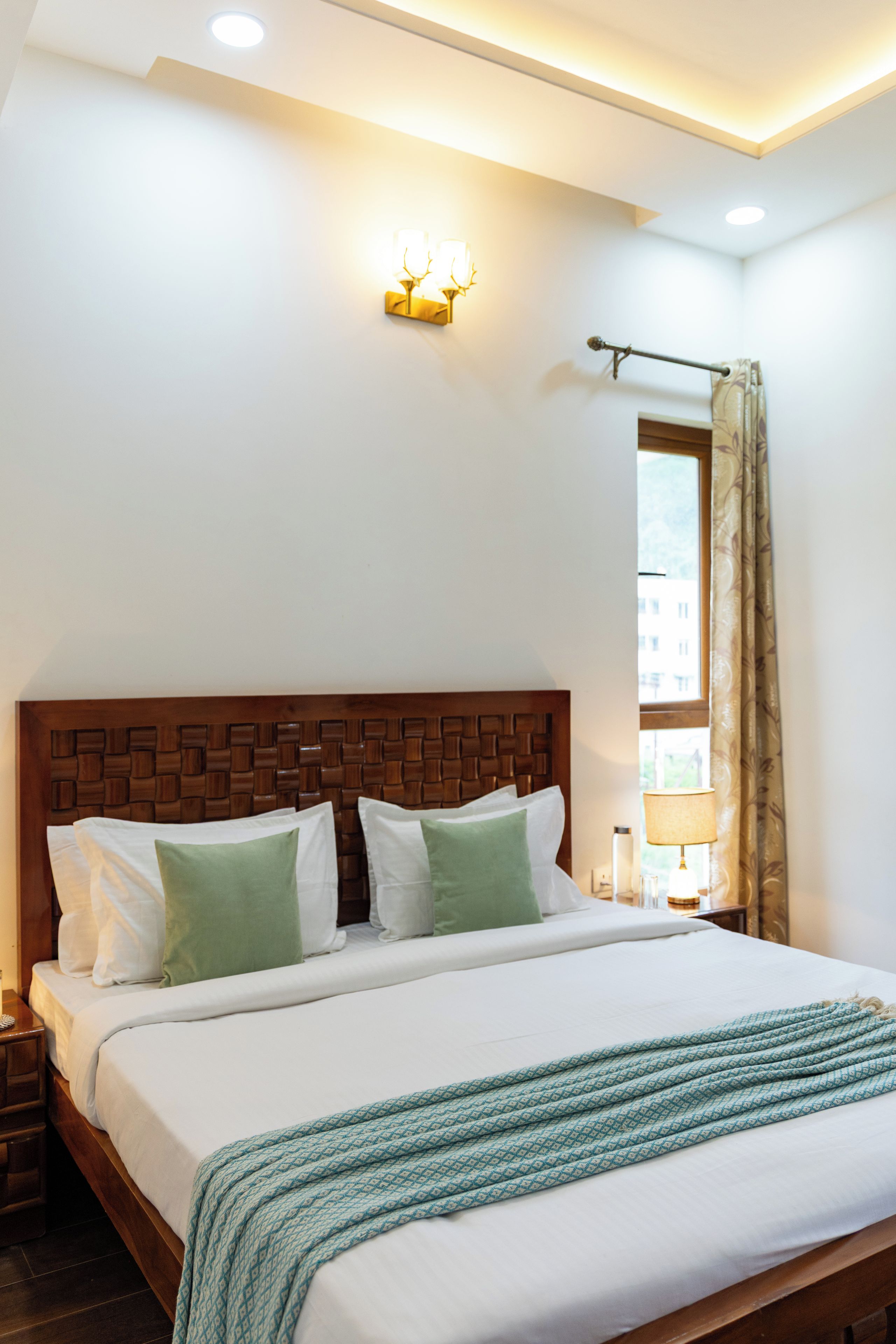 A hotel room with a large bed, a wooden headboard, a nightstand, a lamp, and a window with curtains.