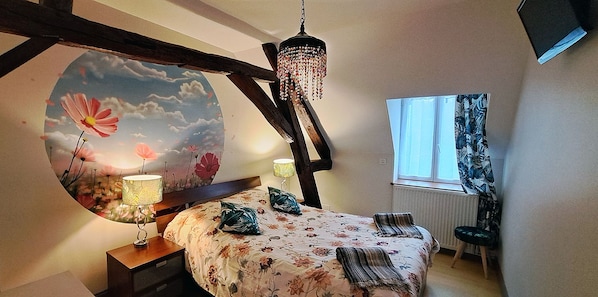 A bedroom with a bed, two bedside lamps, a hanging chandelier, a window with curtains, and a wall mural.