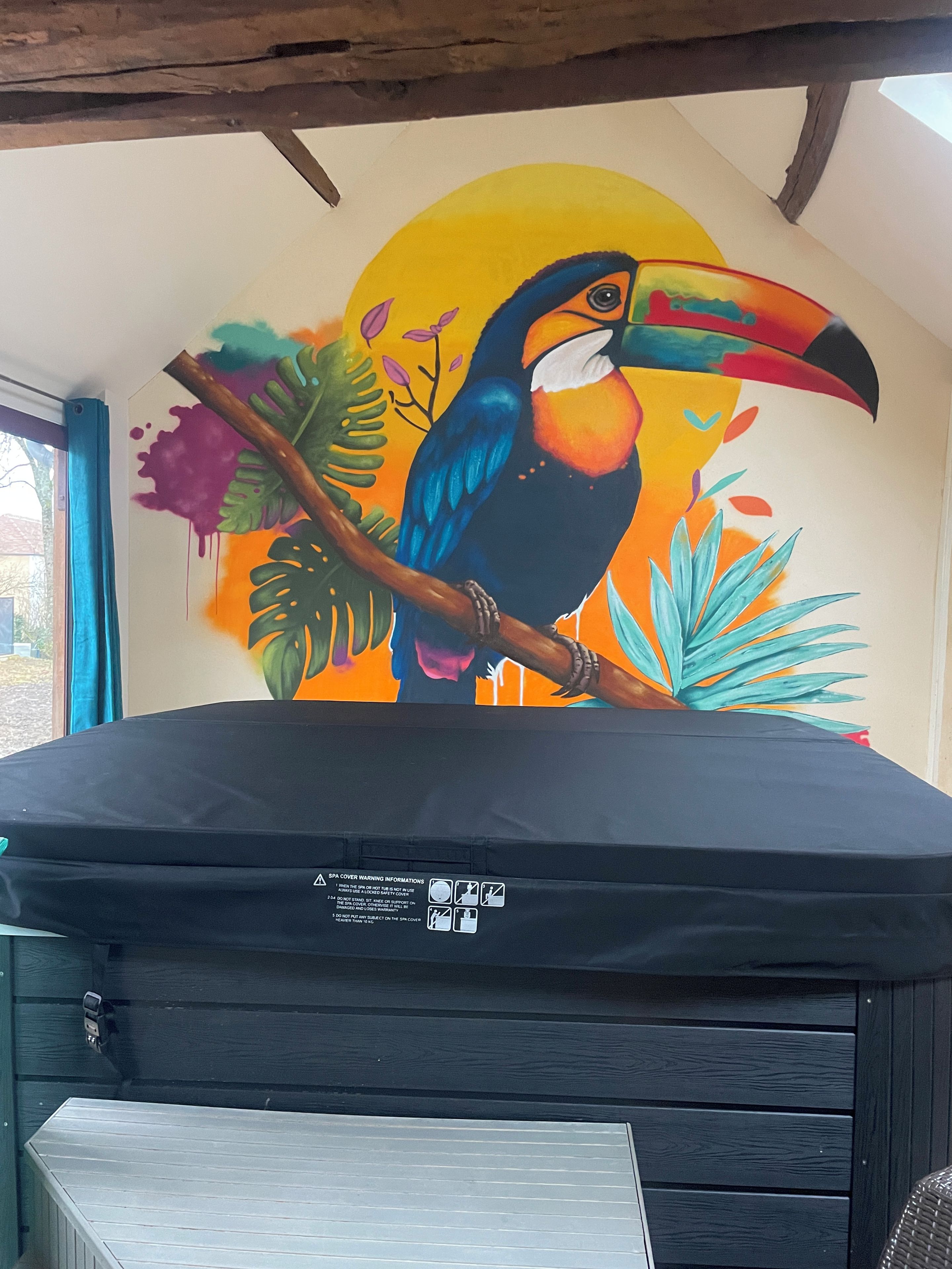 A room with a mural of a toucan on the wall, a window with blue curtains, and a black sofa.