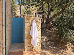 A stone wall with a blue tiled shower area, a white towel hanging on the wall, and a natural outdoor setting with trees and rocks. - Romantic designed stone house with pool & views (Palau-Saverdera)