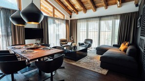 Ein modernes Wohnzimmer mit einem Holzesstisch, Ledersesseln, einem schwarzen Sofa und einem großen Fenster mit Vorhängen.