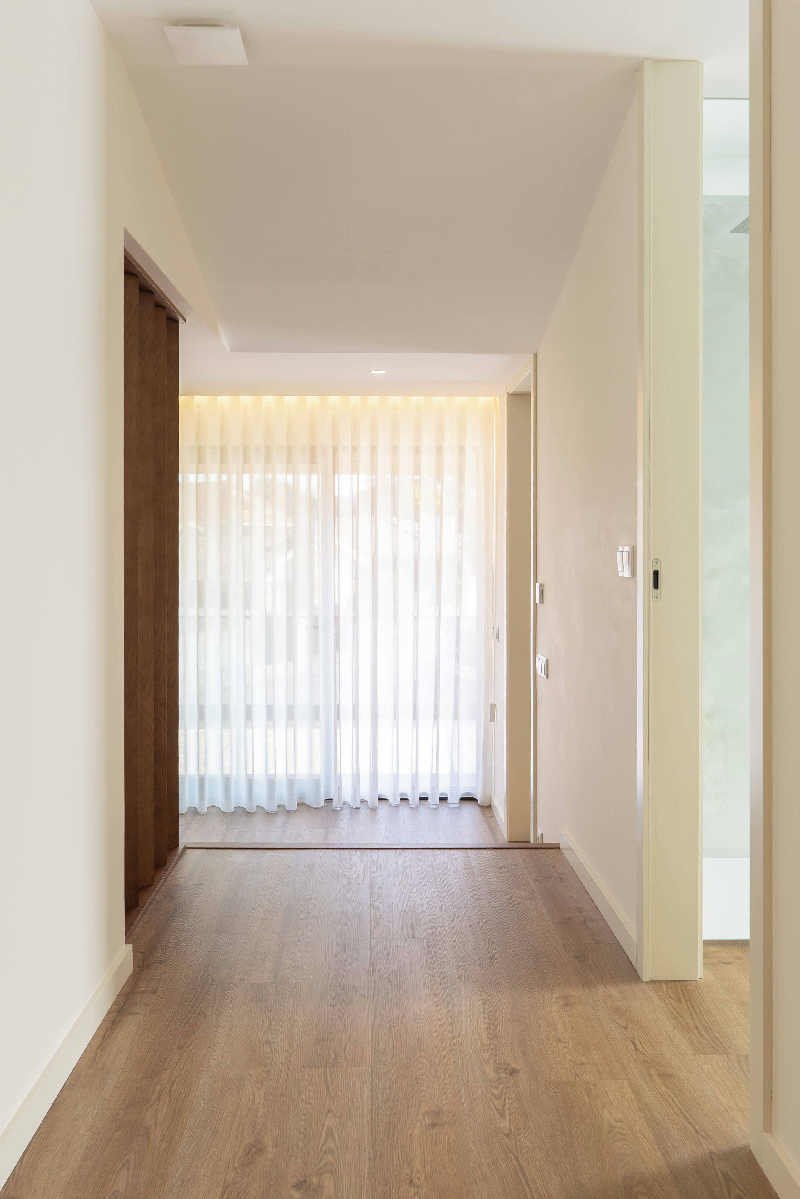 Un pasillo con suelo de madera, una puerta a la derecha y un ventanal con cortinas traslúcidas.