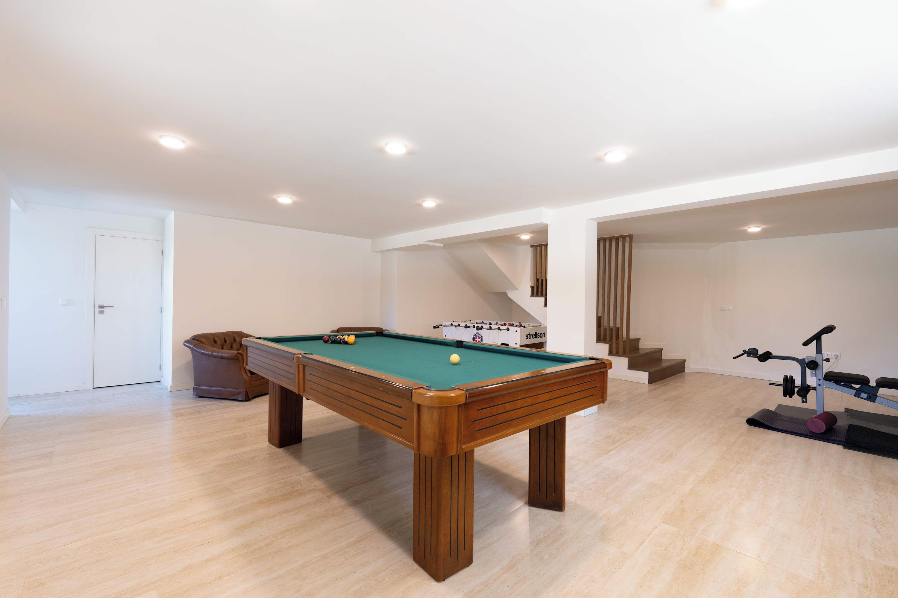 Una habitación amplia con mesa de billar, sofá, cubo de basura y una escalera que sube a otro nivel.