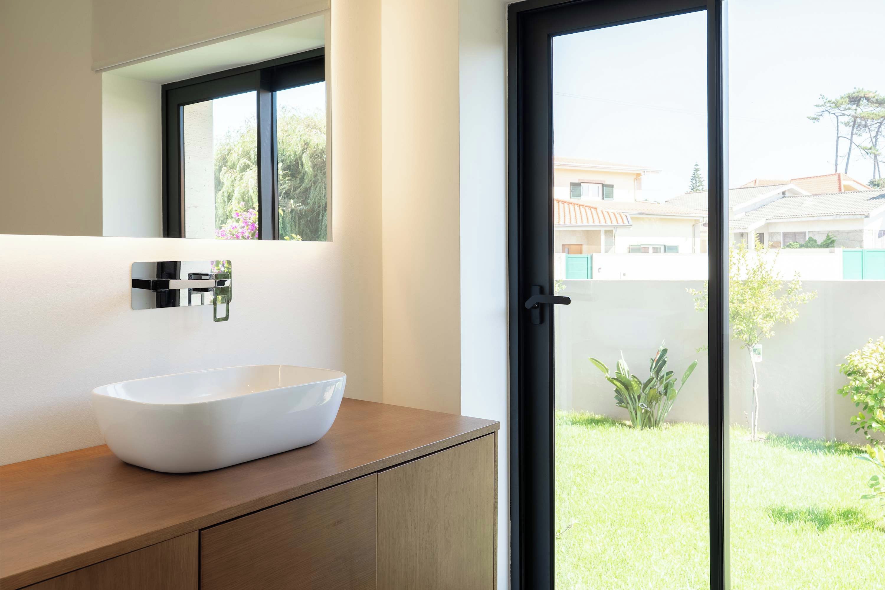 Un baño moderno con un lavamanos blanco tipo recipiente, muebles de madera y un amplio ventanal con vistas a una zona residencial.