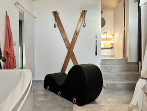 Un bagno moderno con una sedia nera, un portasciugamani in legno a forma di X e una scala a vista che conduce a una camera da letto.