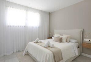Ein Schlafzimmer mit einem Bett, weißen Vorhängen, einem Nachttisch mit Lampe und einem Wandregal.