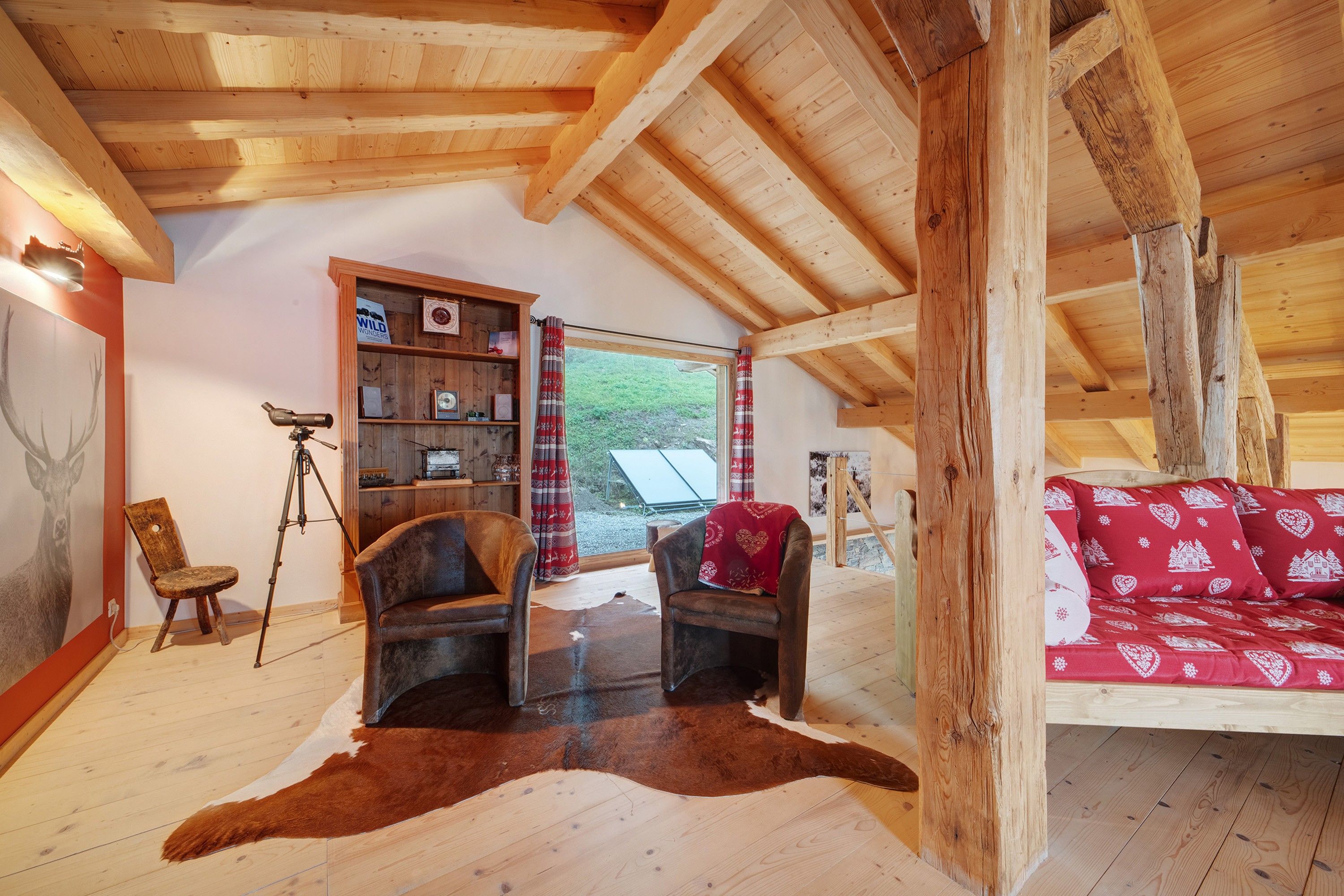 Une pièce chaleureuse avec des poutres en bois, une bibliothèque, un bureau avec un télescope, un fauteuil en cuir, un tapis en peau de vache et un canapé rouge à motifs.