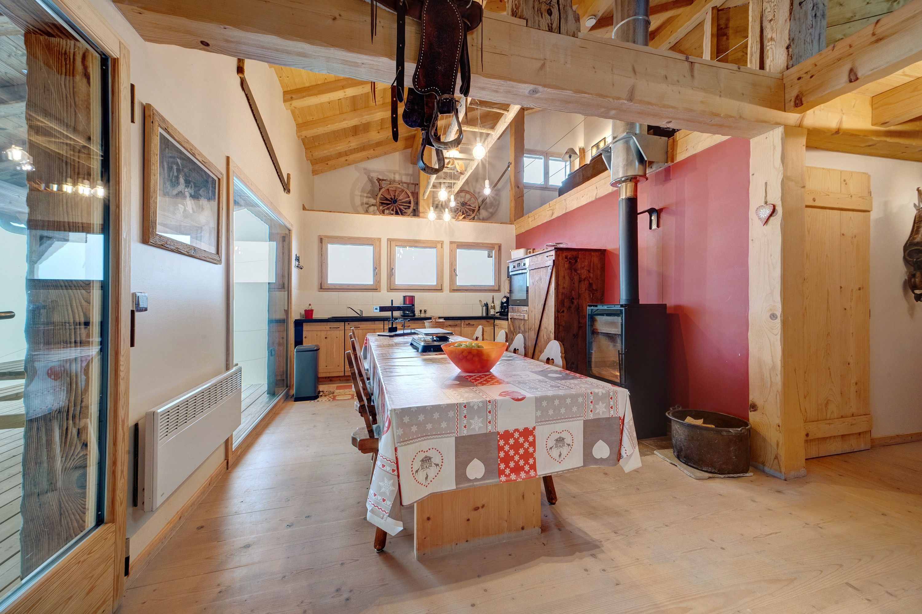 A spacious kitchen with a long wooden table, a red wall, and a rustic ceiling.