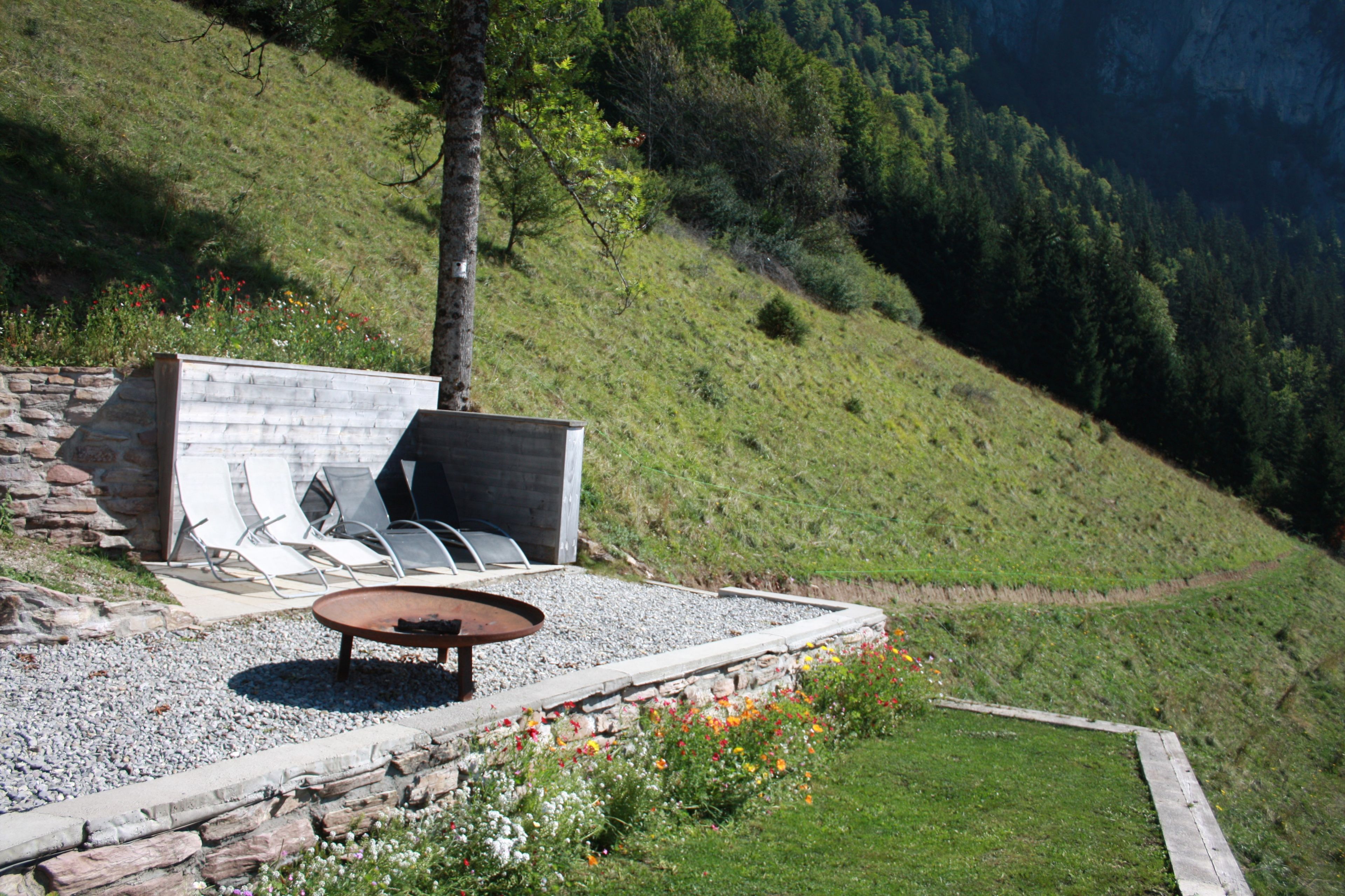 Un área de estar al aire libre con un banco de piedra y una mesa, rodeada por un camino de grava y un parterre floral.