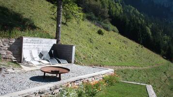 Un área de estar al aire libre con un banco de piedra y una mesa, rodeada por un camino de grava y un parterre floral.