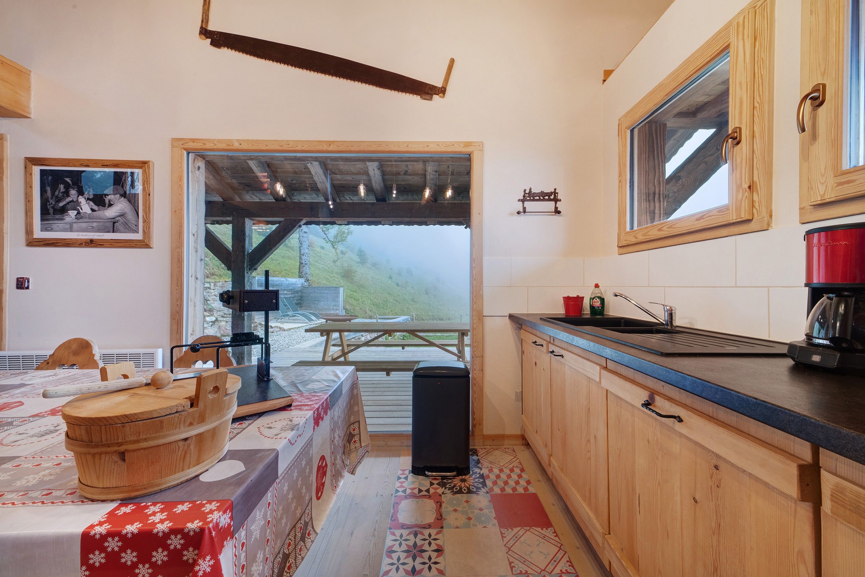 Una cocina rústica con armarios de madera, una mesa de comedor lista para dos personas y vistas a un área exterior por una puerta abierta.