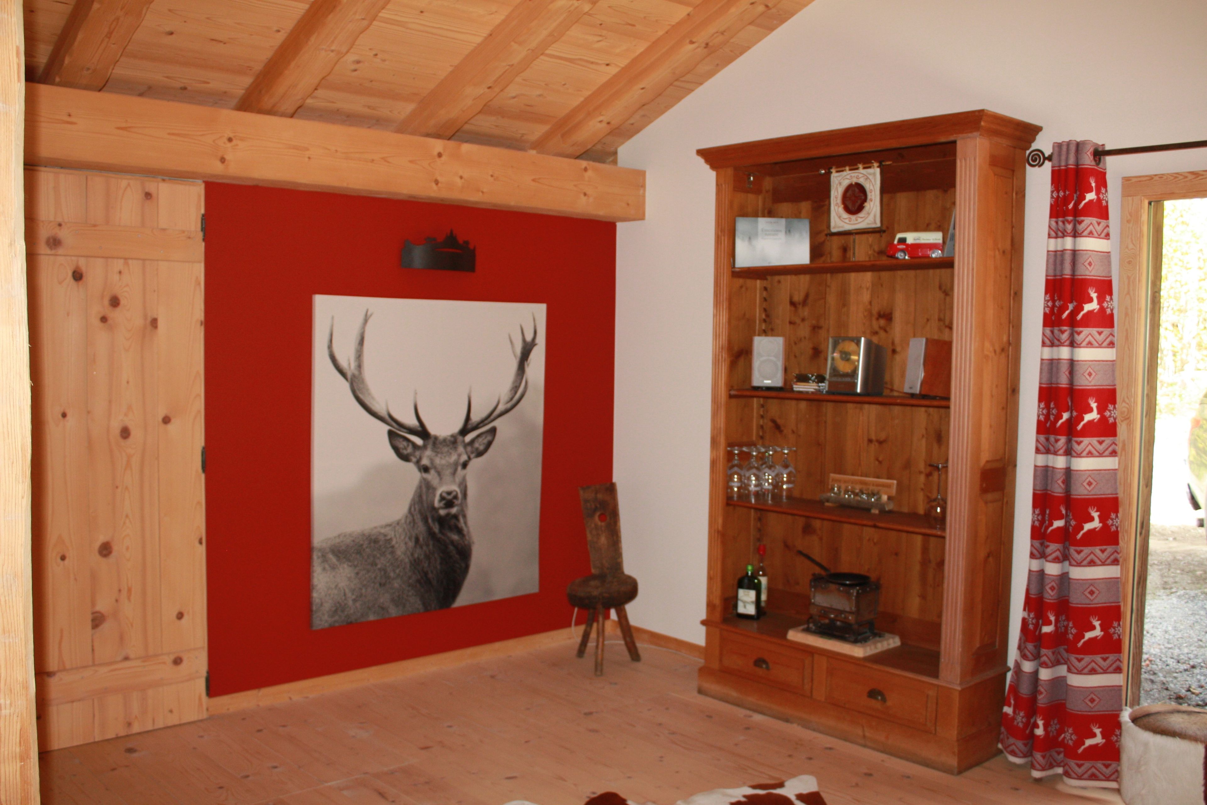 Una habitación con paredes de madera, un cuadro grande de un ciervo enmarco, un mueble de madera con cristalería y una cortina roja.