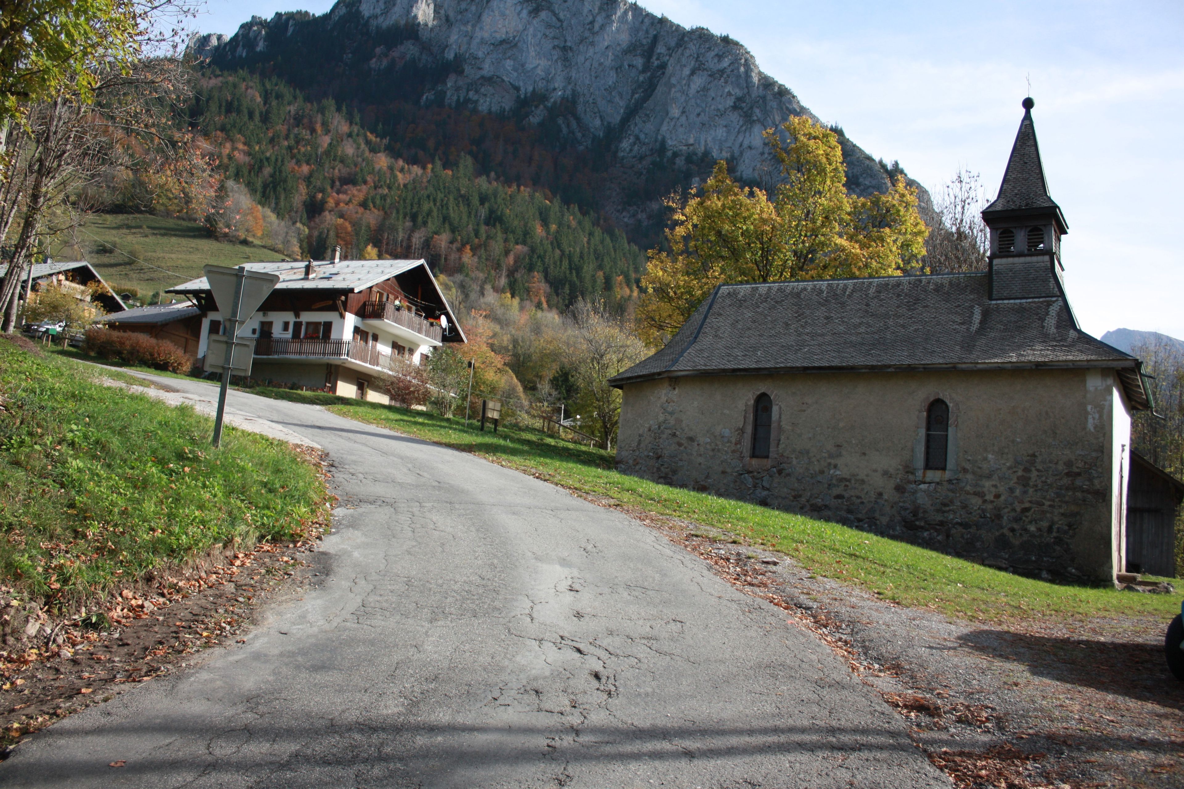 A rural road leading to a stone church with a bell tower, surrounded by autumnal trees and a mountain backdrop.