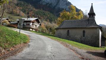 A rural road leading to a stone church with a bell tower, surrounded by autumnal trees and a mountain backdrop.