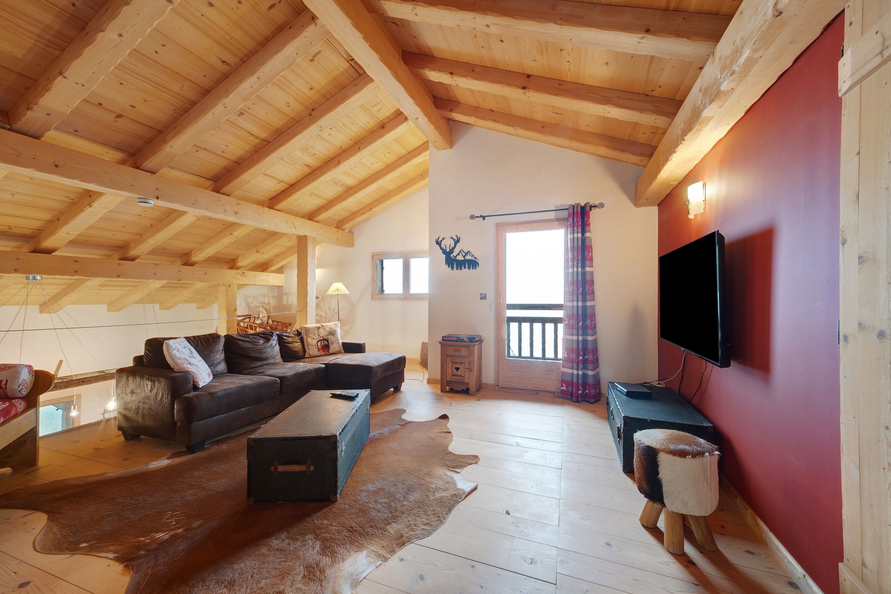 A spacious loft with a high wooden ceiling, a cozy seating area with a sofa and a coffee table, a television mounted on the wall, and a large window with a view.