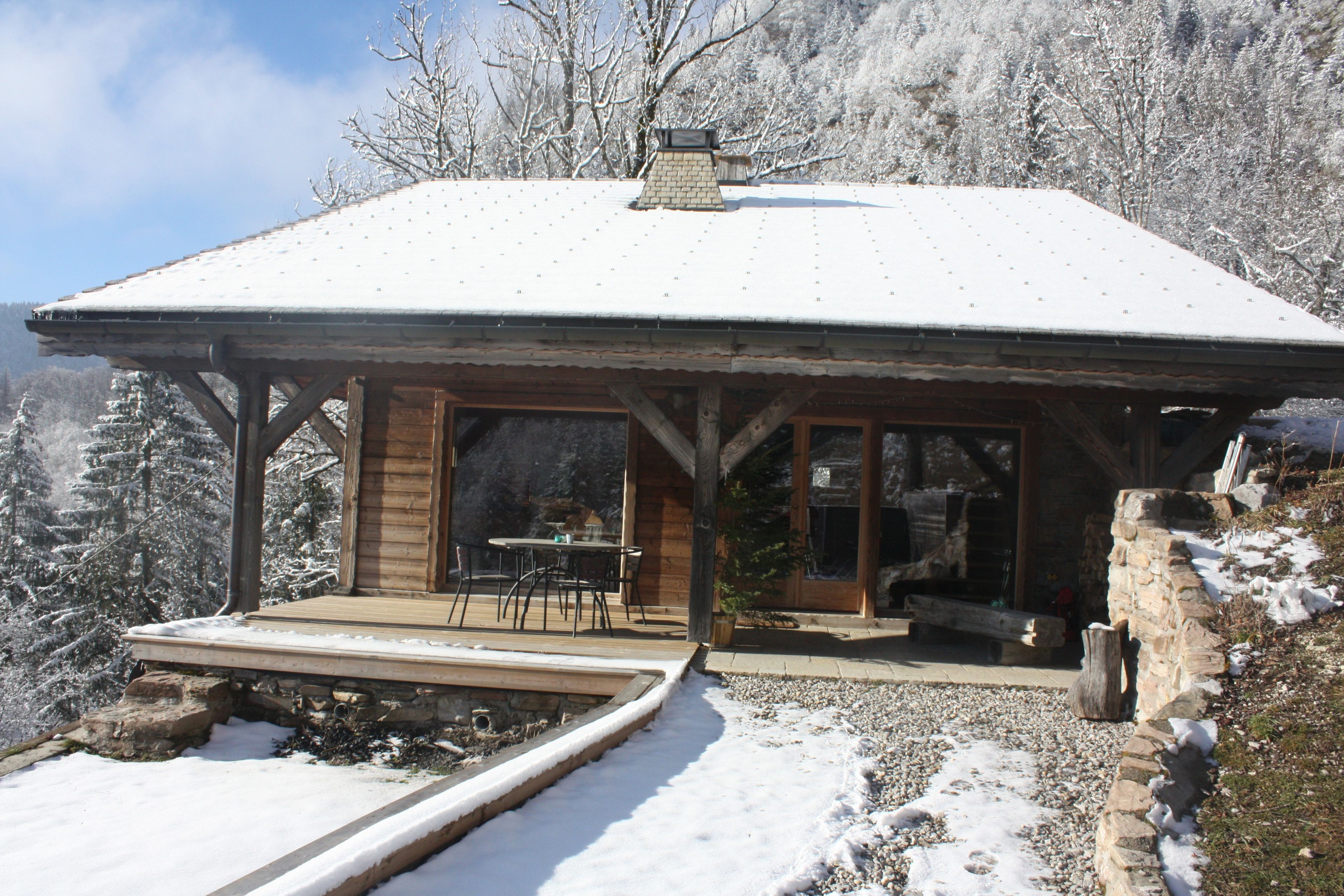 Une cabane en bois avec un toit enneigé, une terrasse avec une table et des chaises, et une vue sur des arbres couverts de neige.