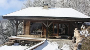 A wooden cabin with a snow-covered roof, a patio with a table and chairs, and a view of snow-covered trees.