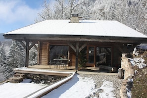 A wooden cabin with a snow-covered roof, a patio with a table and chairs, and a view of snow-covered trees.