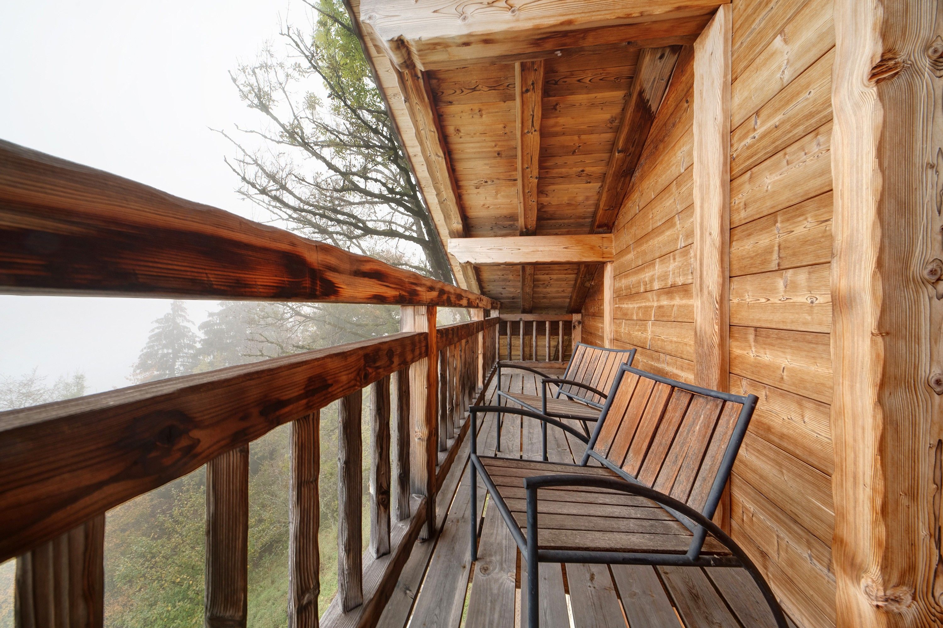 Un balcón de madera con barandillas metálicas y piso de madera, con vista a un área boscosa.