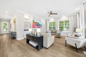 A spacious living room with a flat-screen TV, a ceiling fan, and a large window with curtains. - NEW! Cozy Front Porch, Playroom, Firepit, Kayaks! (Mooresville)