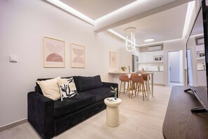 A modern living room with a black sofa, a small round table, and a kitchen area in the background. - FiftySeven Athens - Lemon Garden Hideaway | B3 (Athina)