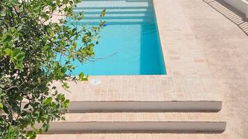 Una piscina rectangular con superficie de agua azul cristalina, rodeada por una solana con baldosas y una pared blanca.