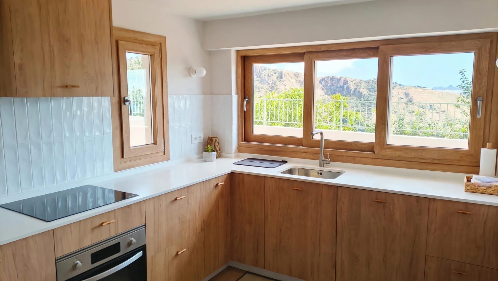 Una cocina con armarios de madera, encimera blanca, horno empotrado, un lavabo y una ventana con vistas a zonas verdes y montañas.