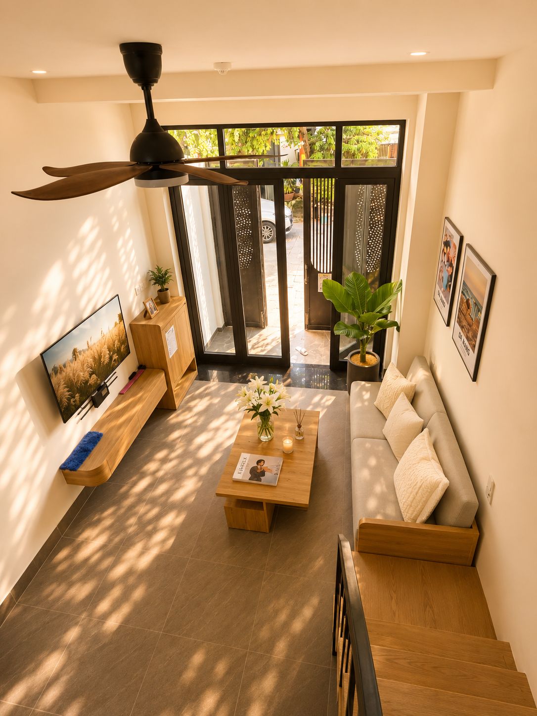 A modern living room with a sofa, coffee table, and a ceiling fan. There is a TV mounted on the wall, a small table with a vase of flowers, and a staircase leading to another level.