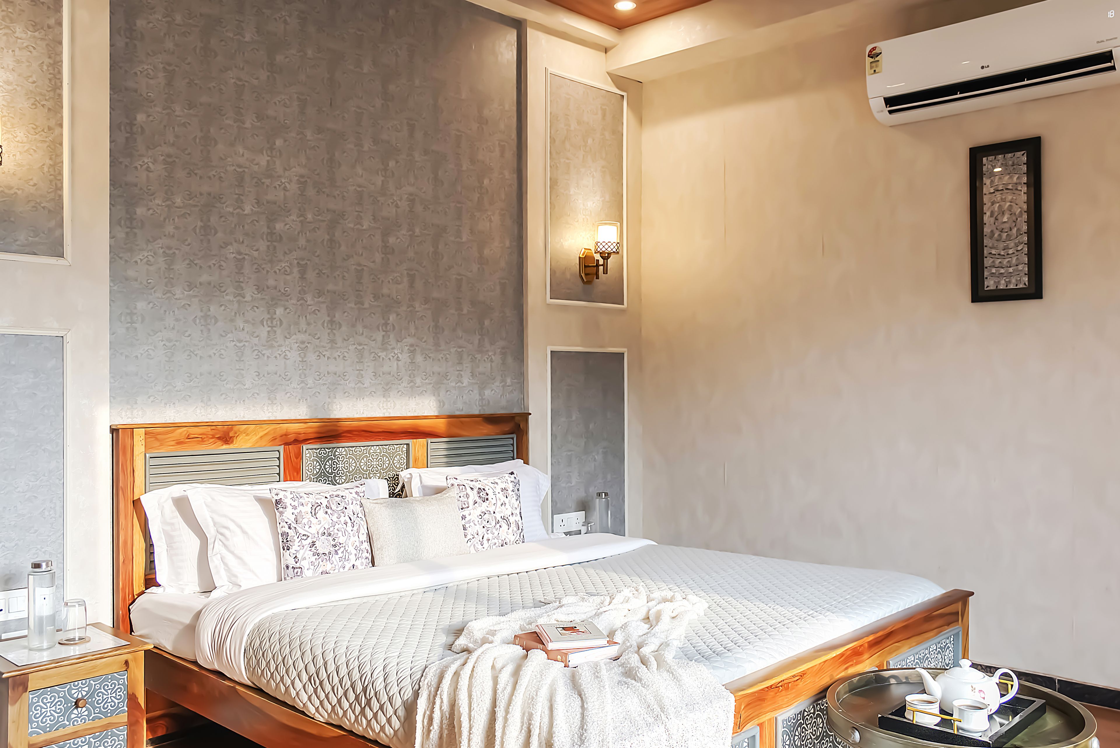 Een net opgemaakt bed met een gewatteerde hoes, een nachtkastje met een glazen vaasje, en een wandairconditioning.