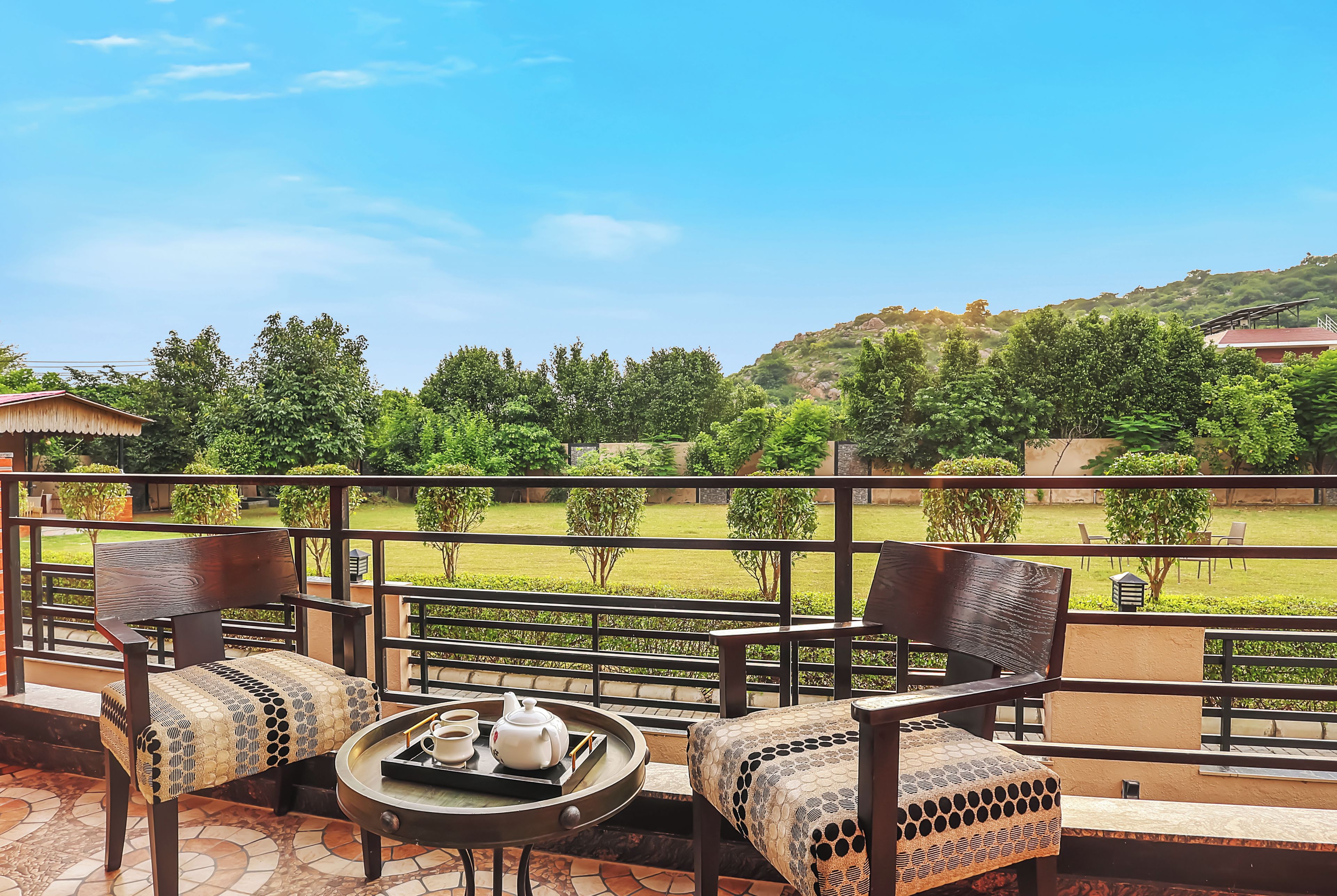 A terrace with a table set for tea, chairs, and a view of a landscaped garden and hills.