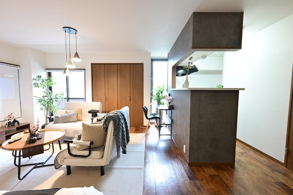A modern living room with a sofa, a coffee table, and a dining area with a small table and chairs. There is a wooden cabinet and a plant on the right side.