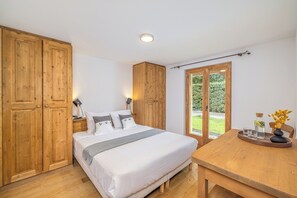 Een slaapkamer met een bed, een houten garderobe, bedlampjes, een tafel met een vaas en uitzicht door een glazen deur.