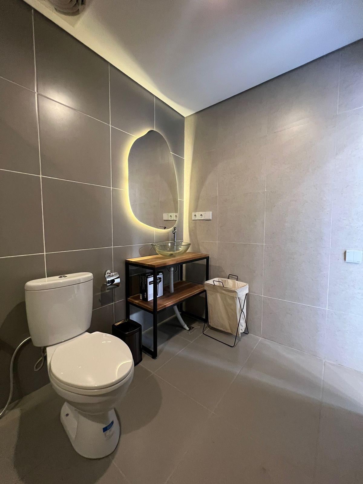 A modern bathroom with a toilet, a wooden vanity with a glass bowl sink, and a mirror with integrated lighting.