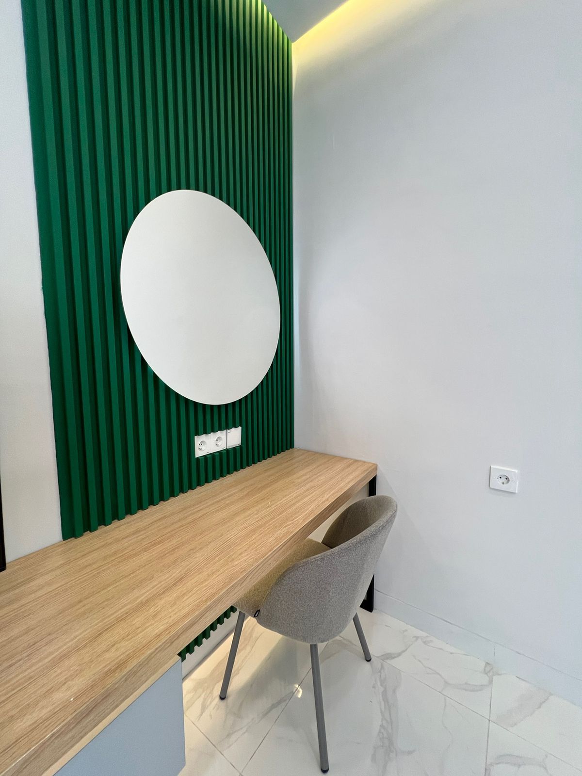 A reception desk with a wooden surface, a grey chair, a green vertical panel, and a white circular mirror.