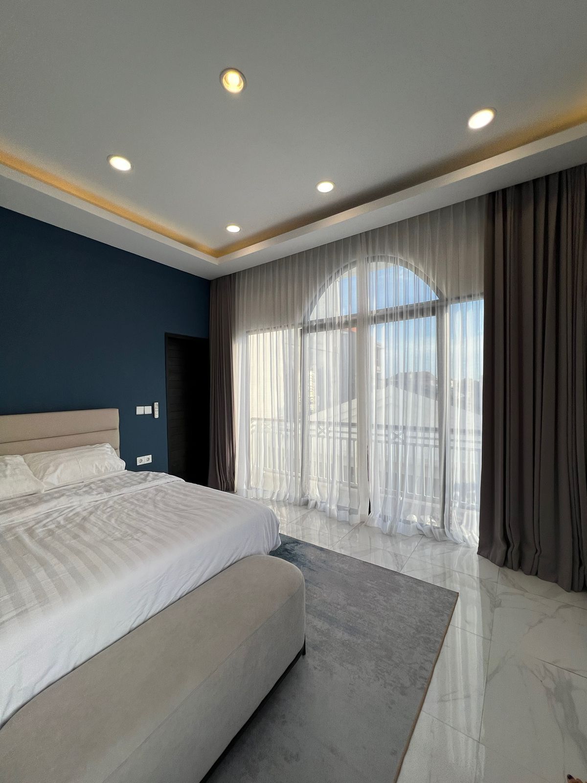 Un dormitorio moderno con una cama grande, vistas de la ciudad a través de cortinas translúcidas e iluminación empotrada en el techo.