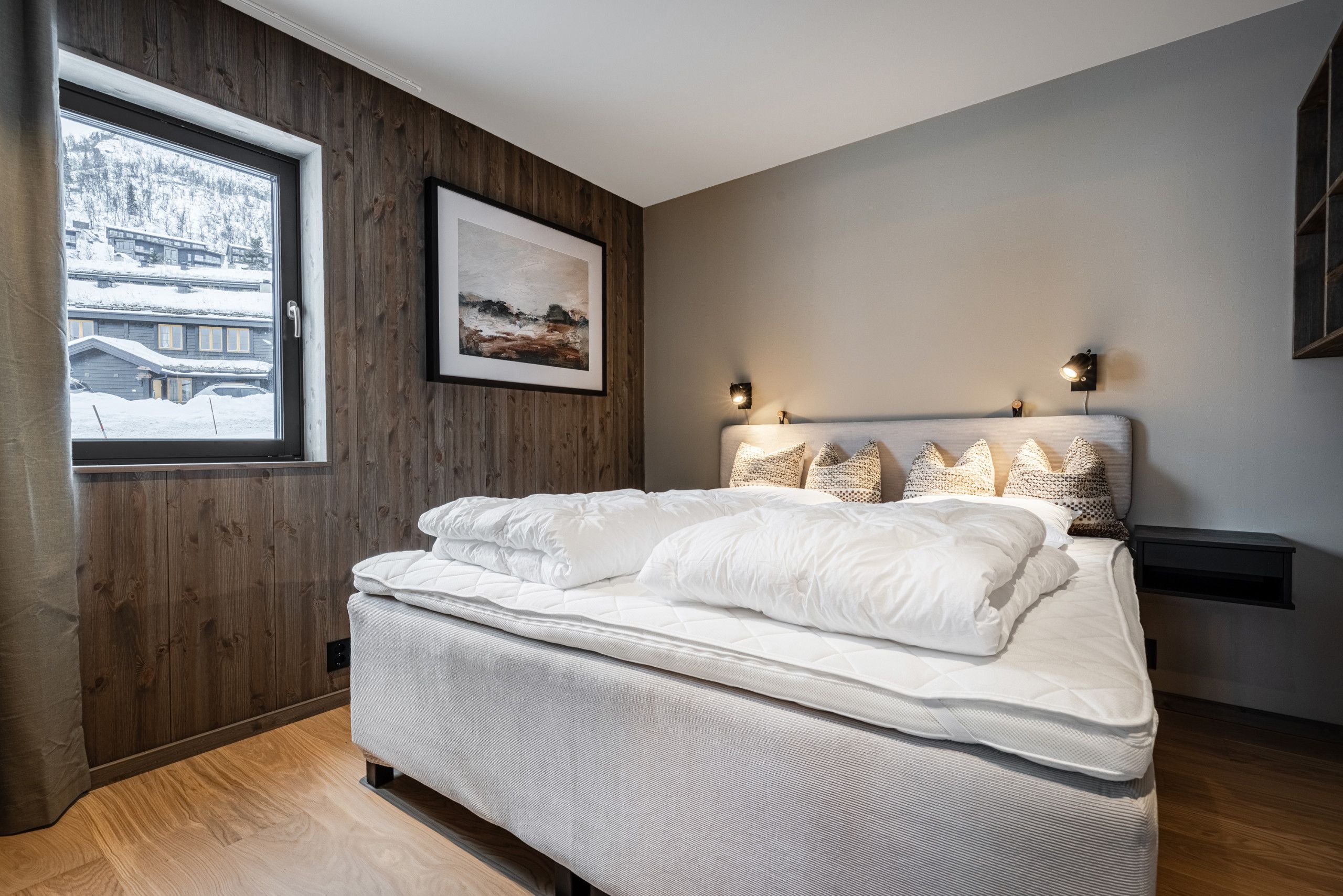 Une chambre avec un mur en bois, un lit avec une literie blanche, un tableau encadré et deux lampes fixées au mur.