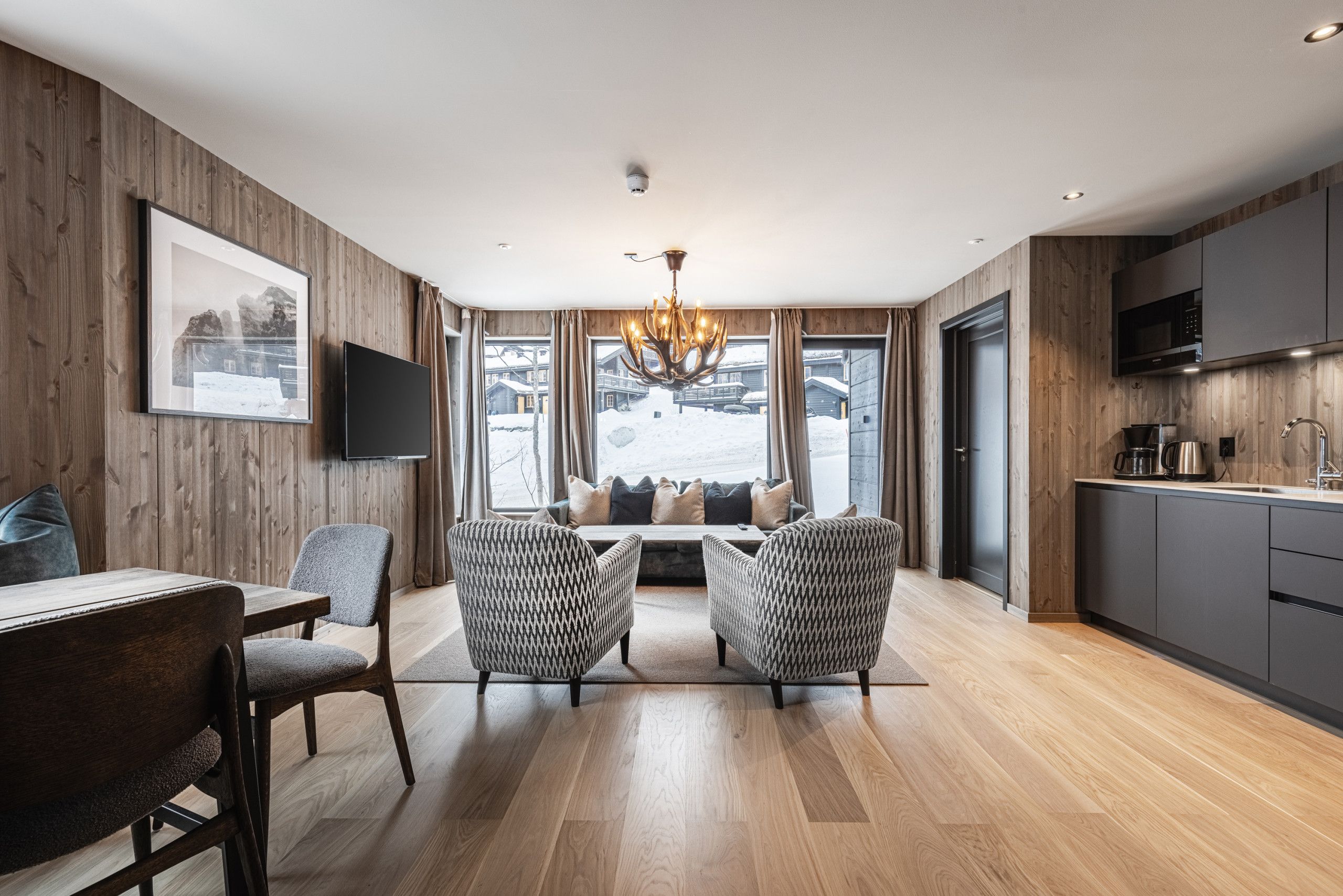 A modern living room with a wooden floor, a dining area with a table and chairs, a large window with a view of a snowy landscape, and a kitchen with a flat-screen TV.