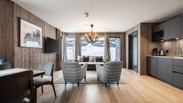 A modern living room with a wooden floor, a dining area with a table and chairs, a large window with a view of a snowy landscape, and a kitchen with a flat-screen TV.