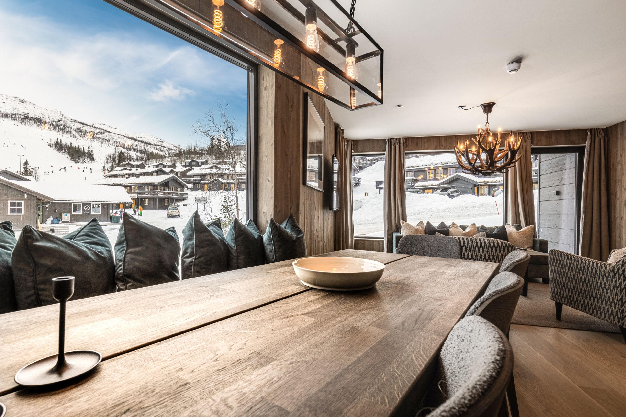 Un intérieur moderne doté d’une grande fenêtre offrant une vue sur les montagnes enneigées, d’un coin repas avec une table en bois et d’un lustre.