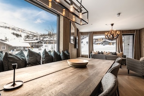 Un intérieur moderne doté d’une grande fenêtre offrant une vue sur les montagnes enneigées, d’un coin repas avec une table en bois et d’un lustre.